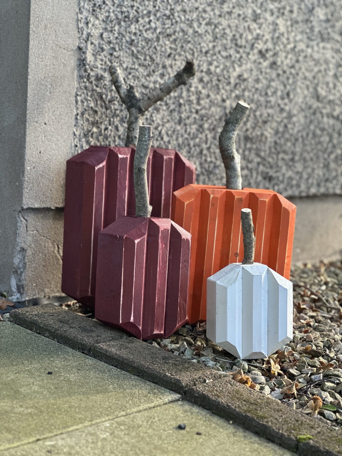 Burgundy, orange and white wooden pumpkins on gravel against a stone wall.