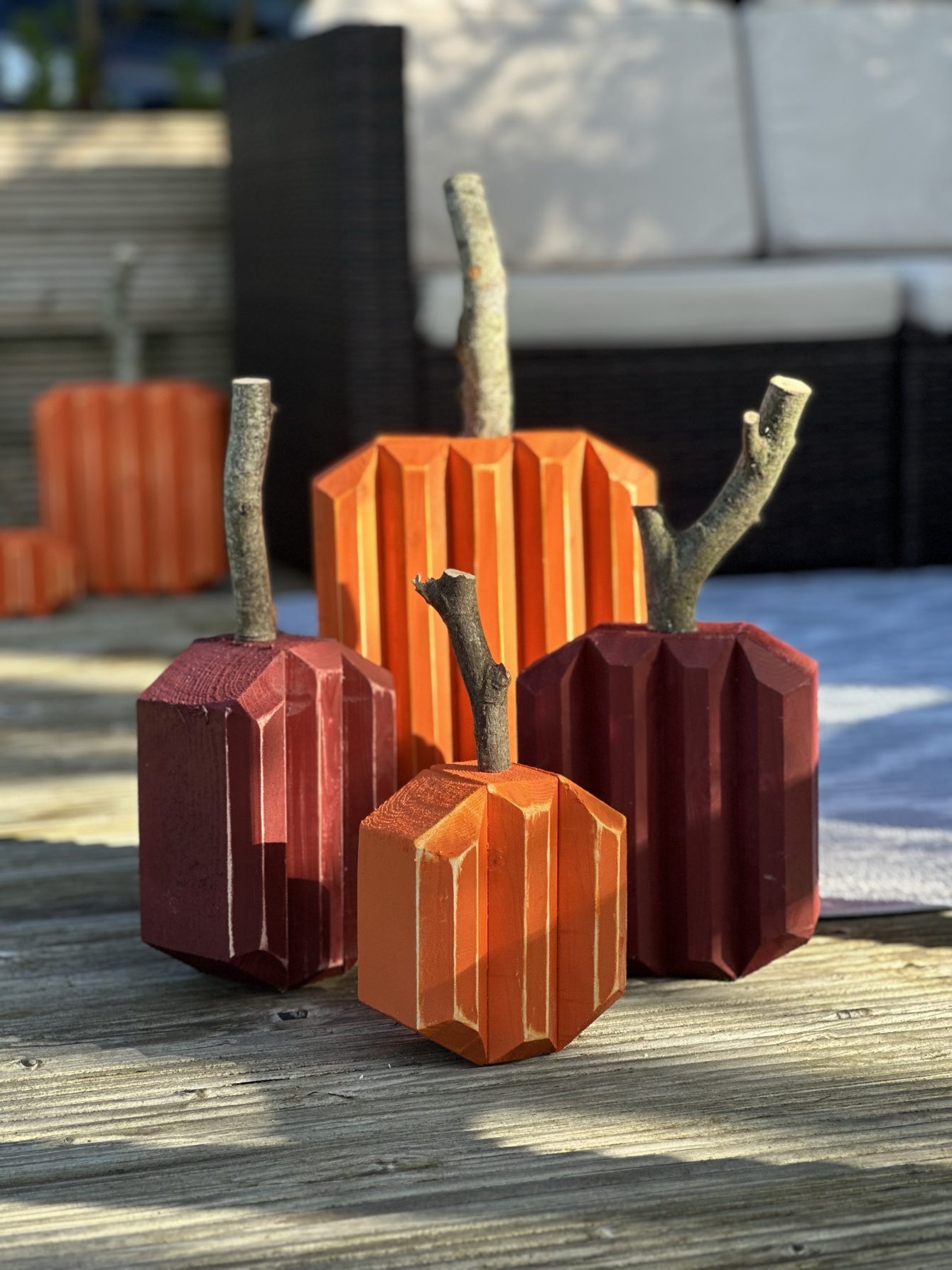 Four wooden pumpkins — two orange, two burgundy — with real branch stems, grouped on a garden deck in low sun.